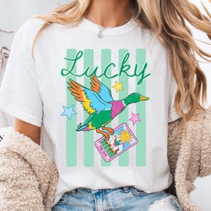 May include: White t-shirt with a colorful duck graphic holding a tarot card that reads "The Sun." The word "Lucky" is written in green script above the duck. Green vertical stripes are behind the duck.