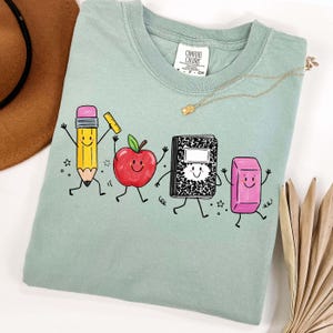 May include: A light green t-shirt featuring a playful illustration of school supplies. The design includes a smiling pencil, apple, notebook, and eraser, all with animated faces and arms. The shirt has a crew neck and short sleeves.