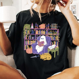 May include: Black t-shirt with a fantasy-themed illustration. A ghost reads in a purple armchair, surrounded by a bookshelf, pumpkin, dragon, and sword. The shirt has a loose fit and is made of a soft material.