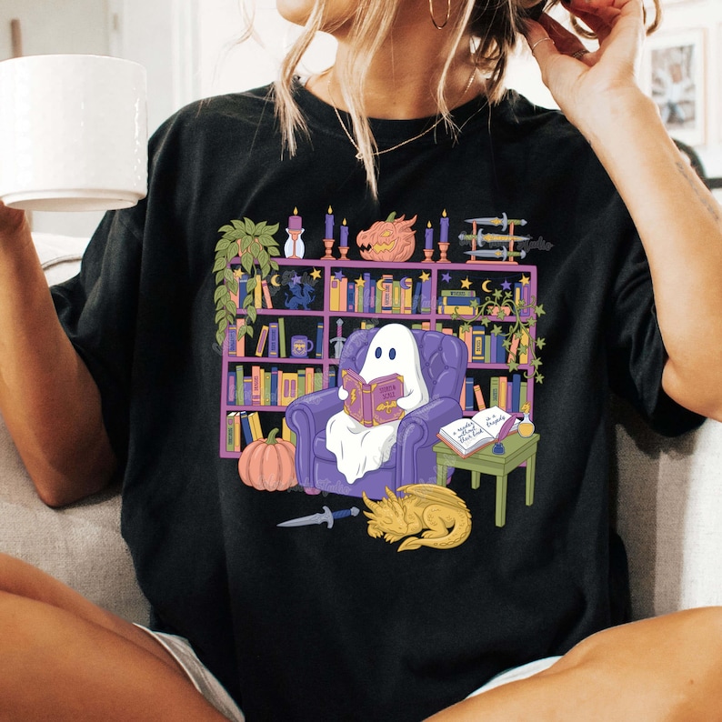 May include: Black t-shirt with a fantasy-themed illustration. A ghost reads in a purple armchair, surrounded by a bookshelf, pumpkin, dragon, and sword. The shirt has a loose fit and is made of a soft material.