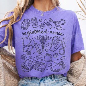 Op de afbeelding: Paars T-shirt met een grafisch ontwerp van medische instrumenten en de tekst "registered nurse".
