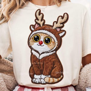 May include: A cream-colored t-shirt featuring an embroidered cartoon cat wearing a reindeer costume. The cat has large, expressive eyes, a red nose, and brown antlers. The cat is sitting, and the costume is brown with white trim.