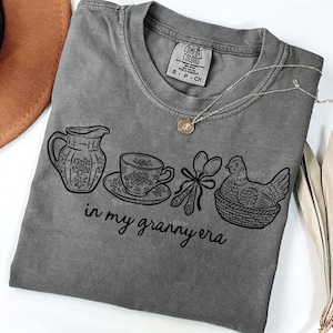 Puede incluir: Una camiseta gris Comfort Colors con un dise&ntilde;o gr&aacute;fico negro que muestra una jarra, una taza de t&eacute;, cucharas y una gallina en una cesta. El texto "in my granny era" est&aacute; debajo. Se ve un collar dorado con un colgante.