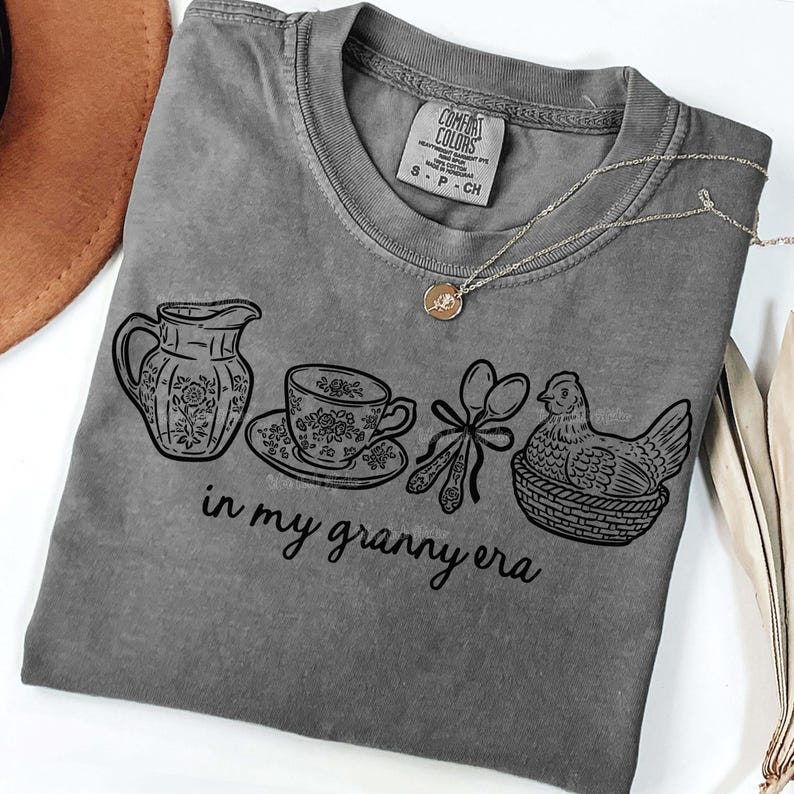 Puede incluir: Una camiseta gris Comfort Colors con un dise&ntilde;o gr&aacute;fico negro que muestra una jarra, una taza de t&eacute;, cucharas y una gallina en una cesta. El texto "in my granny era" est&aacute; debajo. Se ve un collar dorado con un colgante.