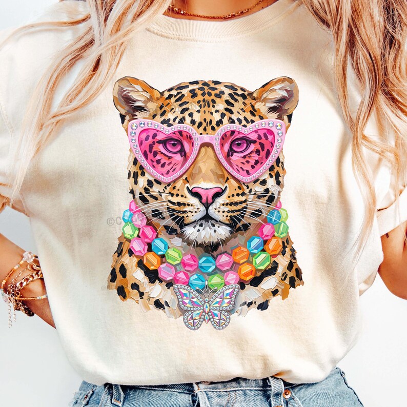 May include: A cream-colored t-shirt featuring a graphic of a leopard wearing pink heart-shaped glasses, a multi-colored beaded necklace, and a butterfly pendant. The leopard has a spotted coat and is centered on the front of the garment.