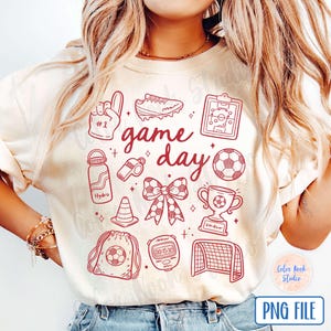 May include: A white t-shirt with a red graphic design that says "game day" and features various sports-related icons, including a football, a whistle, a trophy, a water bottle, a football, and a pair of boots.