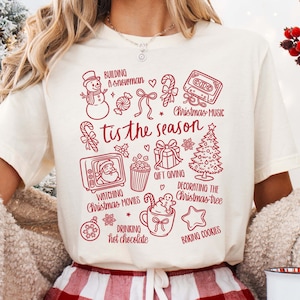 May include: A cream-coloured t-shirt featuring a red Christmas graphic. The design includes a snowman, candy canes, a cassette tape, a Christmas tree, and the phrase "tis the season". Other elements include a TV, popcorn, a gift, a mug, and a star.