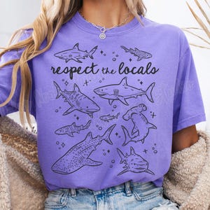 May include: Purple t-shirt with a graphic of various sharks and the text "respect the locals".