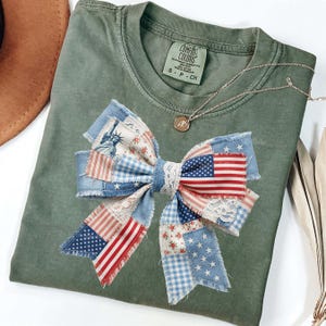 May include: An olive green Comfort Colors t-shirt with a large bow applique. The bow is made of patchwork fabric featuring the American flag, stars, and floral patterns. A silver necklace with a pendant is also visible.