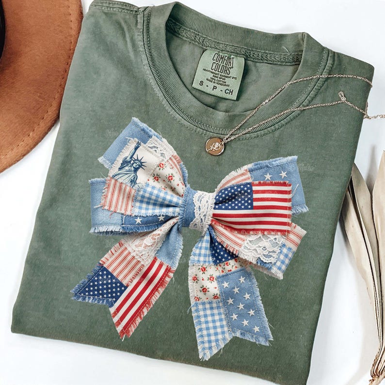May include: An olive green Comfort Colors t-shirt with a large bow applique. The bow is made of patchwork fabric featuring the American flag, stars, and floral patterns. A silver necklace with a pendant is also visible.