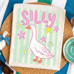 May include: Cream-colored t-shirt with the word "SILLY" in pink letters above a cartoon goose. The goose is white with orange feet and beak, surrounded by pink stars, yellow stars, and daisies. The background has light green vertical stripes.