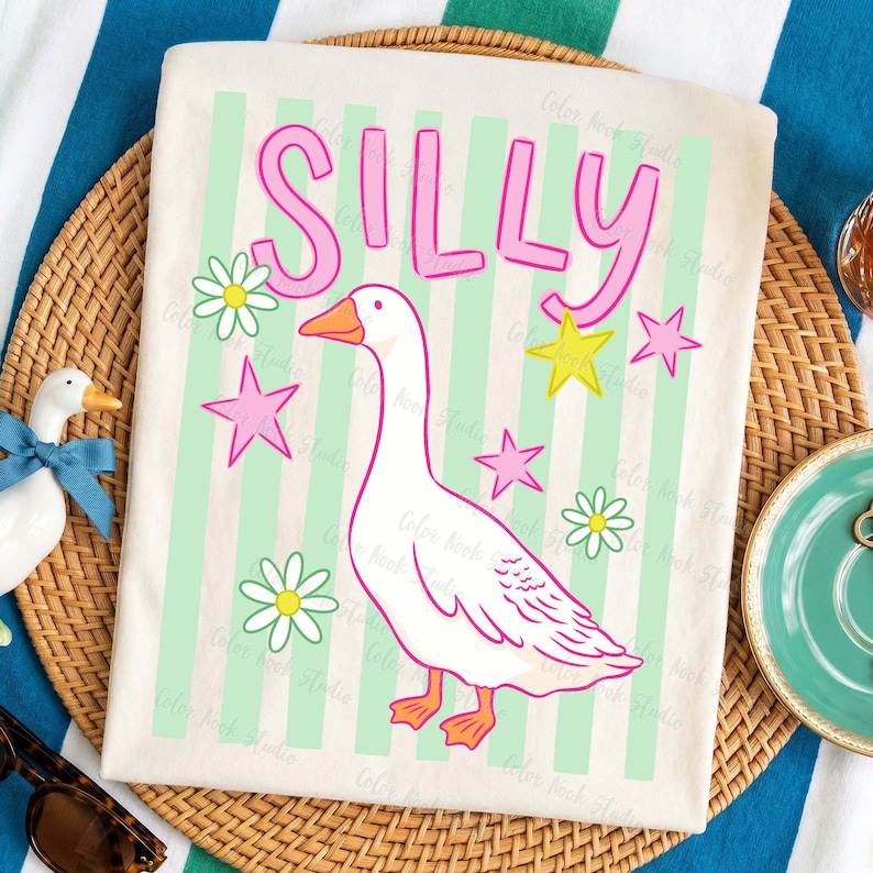 May include: Cream-colored t-shirt with the word "SILLY" in pink letters above a cartoon goose. The goose is white with orange feet and beak, surrounded by pink stars, yellow stars, and daisies. The background has light green vertical stripes.