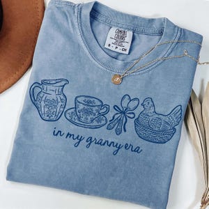 Puede incluir: Camiseta Comfort Colors azul claro con un dise&ntilde;o gr&aacute;fico de una jarra, una taza de t&eacute;, cucharas y una gallina en una cesta. El texto "in my granny era" est&aacute; impreso debajo. Un collar dorado est&aacute; sobre la camiseta.