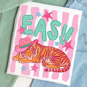 Puede incluir: Camiseta blanca con una ilustraci&oacute;n colorida. El dise&ntilde;o presenta un tigre con un sombrero de vaquero rosa, la palabra "EASY" en letras turquesas y estrellas rosas y azules. El fondo tiene rayas rosas y blancas.
