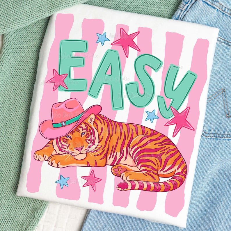 Puede incluir: Camiseta blanca con una ilustraci&oacute;n colorida. El dise&ntilde;o presenta un tigre con un sombrero de vaquero rosa, la palabra "EASY" en letras turquesas y estrellas rosas y azules. El fondo tiene rayas rosas y blancas.