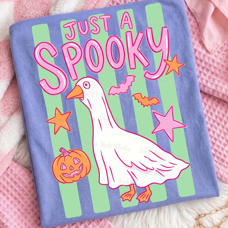 Może przedstawiać: Jasnoniebieska koszulka z r&oacute;żowym napisem "Just a Spooky". Projekt przedstawia kresk&oacute;wkową gęś w kostiumie ducha, pomarańczowe gwiazdy, nietoperze i dynię. Zielone paski biegną pionowo wzdłuż koszulki.
