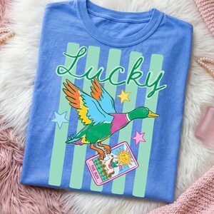 May include: A blue t-shirt featuring a colorful duck with outstretched wings, the word "Lucky" in script, and a tarot card that says "The Sun." The design includes green vertical stripes and star accents.