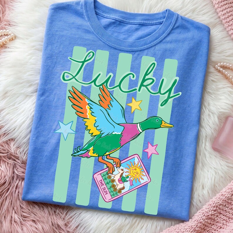 May include: A blue t-shirt featuring a colorful duck with outstretched wings, the word "Lucky" in script, and a tarot card that says "The Sun." The design includes green vertical stripes and star accents.
