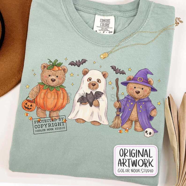 Op de afbeelding: Een saliegroen t-shirt met ronde hals en een afbeelding van drie teddyberen in Halloween-kostuums: &eacute;&eacute;n als pompoen, &eacute;&eacute;n als spook met een vleermuis en &eacute;&eacute;n als tovenaar met een bezem en hoed. De tekst luidt PROTECTED BY COPYRIGHT COLOR NOOK STUDIO.