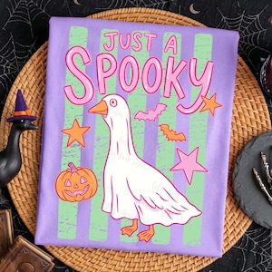 Może przedstawiać: Fioletowa koszulka z napisem "JUST A SPOOKY" w kolorze r&oacute;żowym. Na koszulce znajduje się rysunkowa gęś-duch, pomarańczowe gwiazdy, nietoperze i dynia. Koszulka ma zielone i fioletowe pionowe paski.