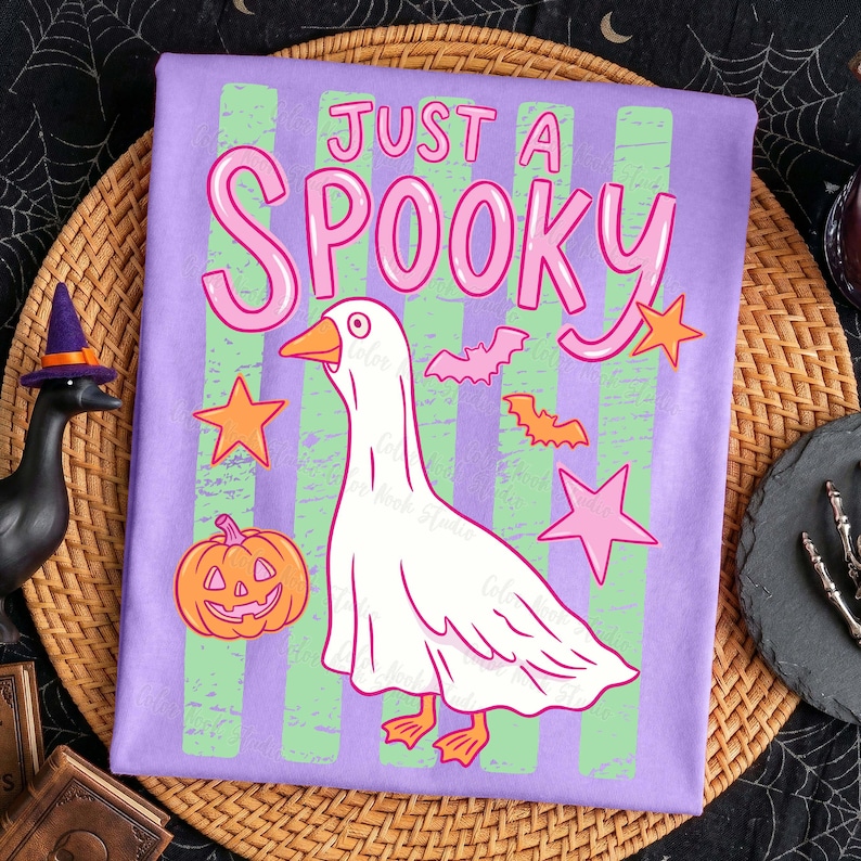 Może przedstawiać: Fioletowa koszulka z napisem "JUST A SPOOKY" w kolorze r&oacute;żowym. Na koszulce znajduje się rysunkowa gęś-duch, pomarańczowe gwiazdy, nietoperze i dynia. Koszulka ma zielone i fioletowe pionowe paski.