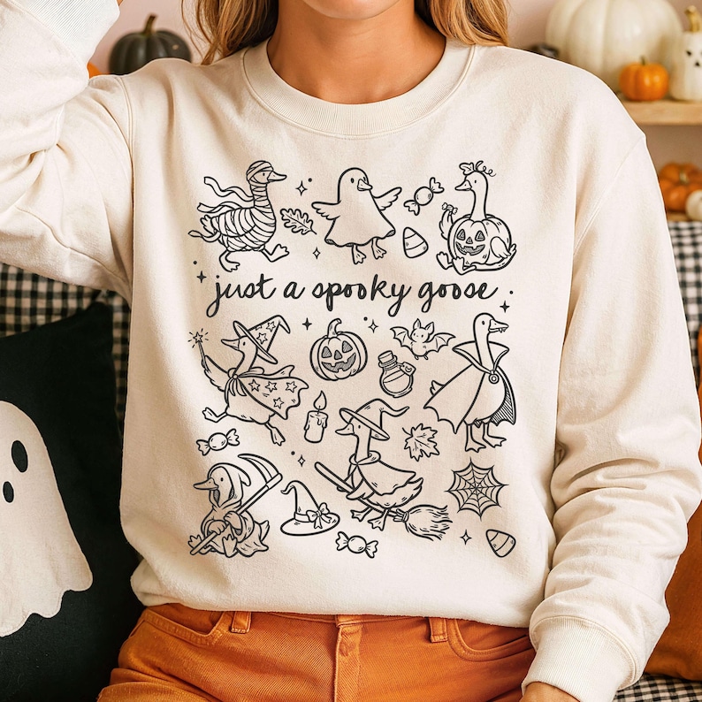 May include: Cream-colored sweatshirt with a black graphic of Halloween-themed geese, pumpkins, and witch hats. The text "just a spooky goose" is printed in a whimsical font. The design is a playful take on Halloween.