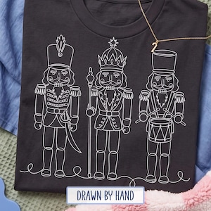 May include: A dark gray t-shirt featuring a white line art graphic of three nutcracker figures. The left figure holds a sword, the center figure holds a staff, and the right figure plays a drum. A thin gold chain necklace rests on the shirt.