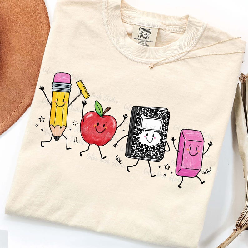 May include: Cream-colored t-shirt featuring a playful graphic of animated school supplies: a pencil, apple, notebook, and eraser. The pencil holds a ruler, and the apple has a green leaf. The shirt has a Comfort Colors tag.
