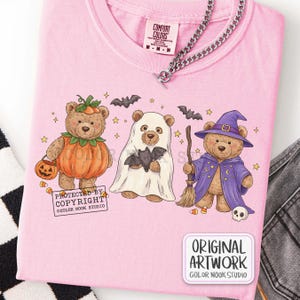 Op de afbeelding: Een lichtroze t-shirt met een afbeelding van drie teddyberen verkleed voor Halloween: &eacute;&eacute;n als pompoen, &eacute;&eacute;n als spook met een vleermuis en &eacute;&eacute;n als tovenaar met een bezem en hoed. Het shirt bevat de tekst PROTECTED BY COPYRIGHT.