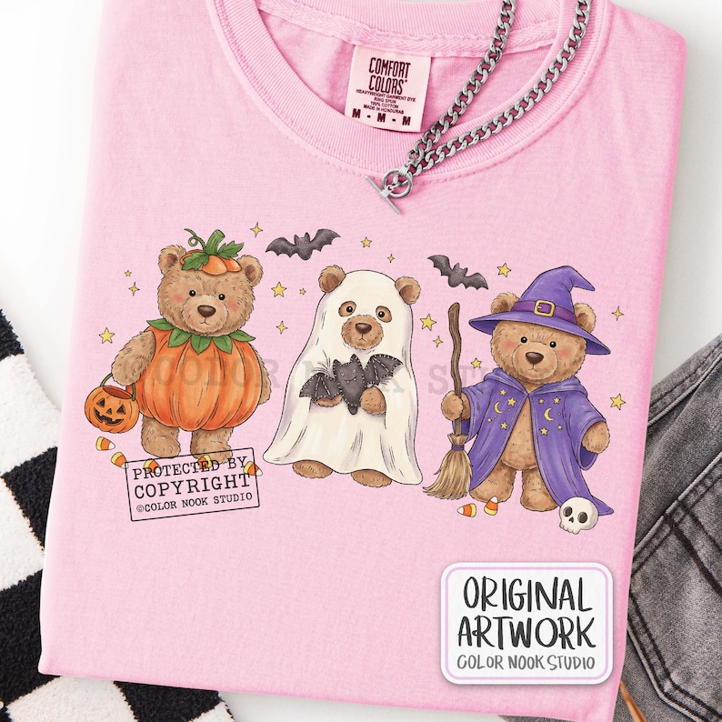 Op de afbeelding: Een lichtroze t-shirt met een afbeelding van drie teddyberen verkleed voor Halloween: &eacute;&eacute;n als pompoen, &eacute;&eacute;n als spook met een vleermuis en &eacute;&eacute;n als tovenaar met een bezem en hoed. Het shirt bevat de tekst PROTECTED BY COPYRIGHT.
