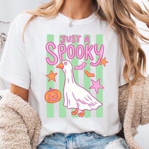 Może przedstawiać: Biała koszulka z grafiką o tematyce Halloween. Projekt zawiera r&oacute;żowy napis "JUST A SPOOKY", kresk&oacute;wkową gęś w kostiumie ducha, nietoperze, gwiazdy i dynię. Tło ma zielone paski.