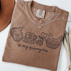 Puede incluir: Una camiseta marr&oacute;n Comfort Colors con un dise&ntilde;o gr&aacute;fico que presenta una jarra, una taza de t&eacute;, cucharas y una gallina en una cesta. El texto "in my granny era" est&aacute; impreso debajo de las ilustraciones. Un collar plateado con un colgante est&aacute; sobre la camiseta.