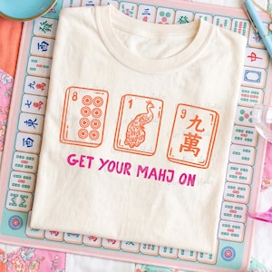 May include: Cream-colored t-shirt with a graphic of three Mahjong tiles in orange and the text "GET YOUR MAHJ ON" in pink. The tiles feature the numbers 8, 1, and 9, with a peacock design on the middle tile. The shirt is on a Mahjong mat.