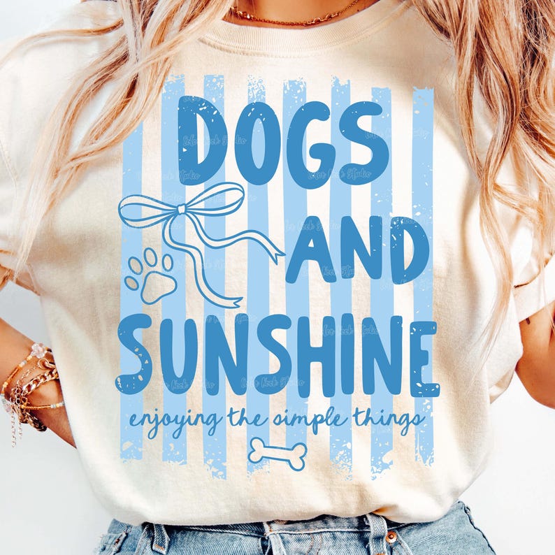 May include: Cream-colored t-shirt with a blue design featuring the words "DOGS AND SUNSHINE" in large letters. The design includes a paw print, bow, and bone, with the phrase "enjoying the simple things" below.