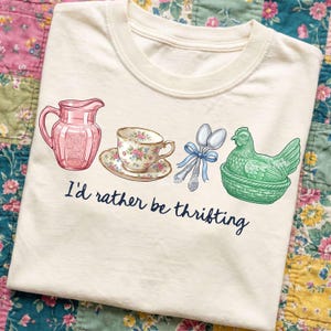 May include: Cream-colored t-shirt featuring a graphic of a pink pitcher, floral teacup, spoons with a blue bow, and a green hen in a basket. The text "I'd rather be thrifting" is printed below the graphics.