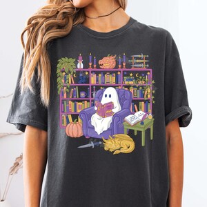 May include: Dark grey t-shirt with a cartoon ghost reading a book in a purple armchair. The ghost is in front of a bookshelf filled with books and decorations. A pumpkin, dragon, and knife are also depicted in the illustration.
