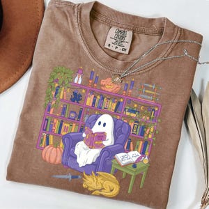 May include: Brown Comfort Colors t-shirt with a quirky illustration of a ghost reading in a purple armchair before a bookshelf. The design includes a pumpkin, candles, and a sleeping dragon. The shirt has a small tag that says "Comfort Colors".