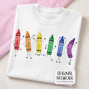 May include: A white t-shirt featuring a graphic of six anthropomorphic crayons in red, orange, yellow, green, blue, and violet. Each crayon has a smiling face, arms, and legs. A small rectangular label in the corner reads ORIGINAL ARTWORK COLOR NOOK STUDIO.