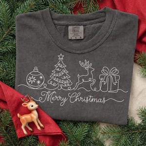 May include: A charcoal grey t-shirt featuring a white continuous line drawing of a Christmas ornament, a pine tree, a reindeer, and a gift box, with the text Merry Christmas written in a cursive script below the graphic elements.