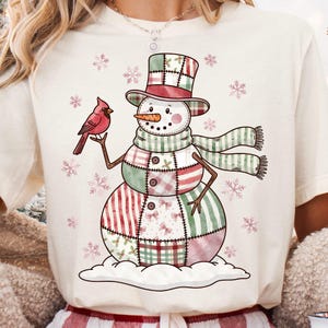 May include: Cream-colored t-shirt featuring a whimsical snowman design. The snowman has a patchwork body in red, green, and white, a carrot nose, and a top hat. A red cardinal bird is perched on its arm, with snowflakes around.