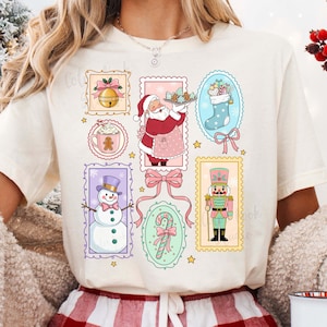 May include: Cream-colored t-shirt featuring a collection of Christmas-themed illustrations. The design includes images of Santa Claus, a snowman, a stocking, a bell, a gingerbread man, and a nutcracker, all within decorative frames.