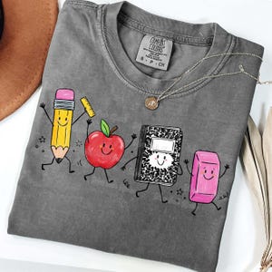 May include: A gray Comfort Colors t-shirt featuring a playful graphic of animated school supplies: a pencil, apple, notebook, and eraser. The pencil holds a ruler, and the apple has a green leaf. A silver necklace with a pendant is also visible.