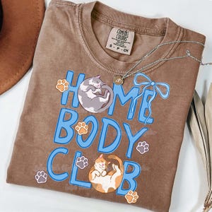 May include: A brown Comfort Colors t-shirt with the words "HOME BODY CLUB" in blue, featuring illustrations of a gray cat, an orange and white cat, and paw prints. A silver necklace and a brown hat are also visible.