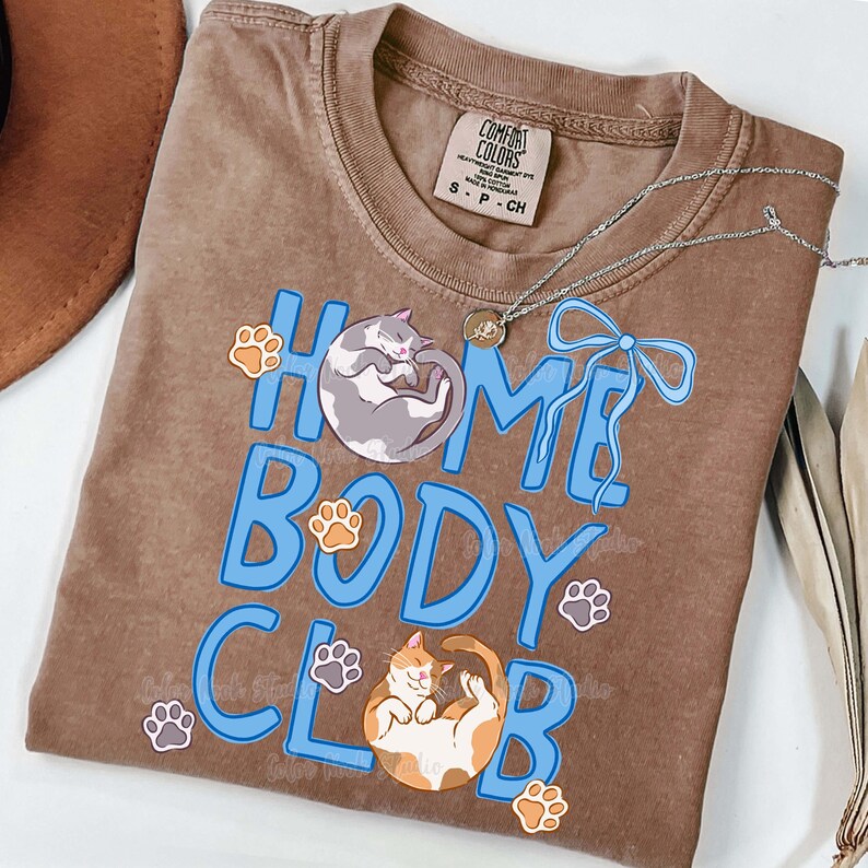 May include: A brown Comfort Colors t-shirt with the words "HOME BODY CLUB" in blue, featuring illustrations of a gray cat, an orange and white cat, and paw prints. A silver necklace and a brown hat are also visible.