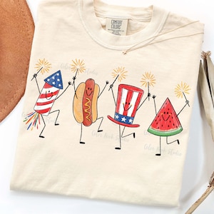 May include: Cream-colored Comfort Colors t-shirt featuring a whimsical illustration of a firework, hot dog, Uncle Sam hat, and watermelon slice, each holding a sparkler. The design has a patriotic theme.