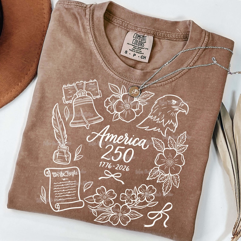 May include: A brown Comfort Colors t-shirt with a white graphic design featuring the Liberty Bell, an eagle, flowers, and the text "America 250 1776-2026". A silver necklace with a small pendant rests on the shirt.