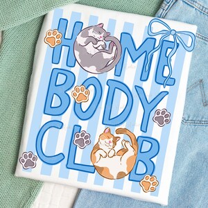 May include: White t-shirt with blue stripes and the words "HOME BODY CLUB" in blue. The shirt features illustrations of two cats and paw prints. One cat is gray and white, curled up, and the other is orange and white, lying on its back.
