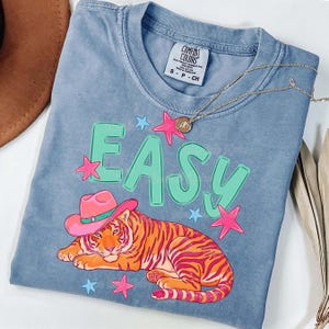 Puede incluir: Camiseta azul Comfort Colors con un tigre con un sombrero de vaquero rosa. La palabra "EASY" est&aacute; impresa en verde, con estrellas rosas y azules. Un collar dorado est&aacute; sobre la camiseta.
