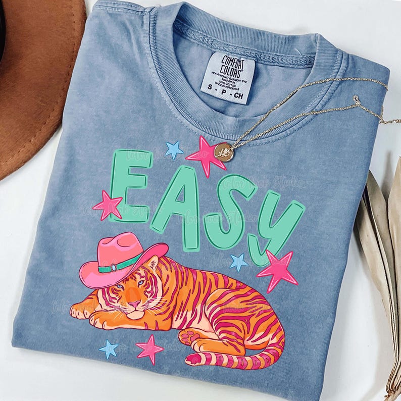 Puede incluir: Camiseta azul Comfort Colors con un tigre con un sombrero de vaquero rosa. La palabra "EASY" est&aacute; impresa en verde, con estrellas rosas y azules. Un collar dorado est&aacute; sobre la camiseta.