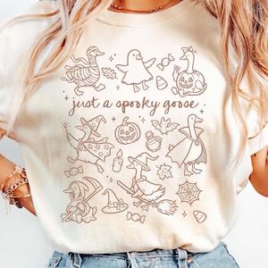 May include: Cream-colored sweatshirt featuring a brown graphic of Halloween-themed geese. The design includes geese dressed as mummies, witches, and vampires, along with pumpkins, candy, and the text "just a spooky goose."